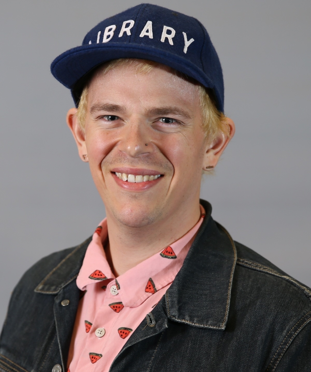 Headshot of Sam Hansen, a white non-binary person with bleach blonde hair, a blue hat reading library, a pink shirt with watermelon on it, and a denim jacket